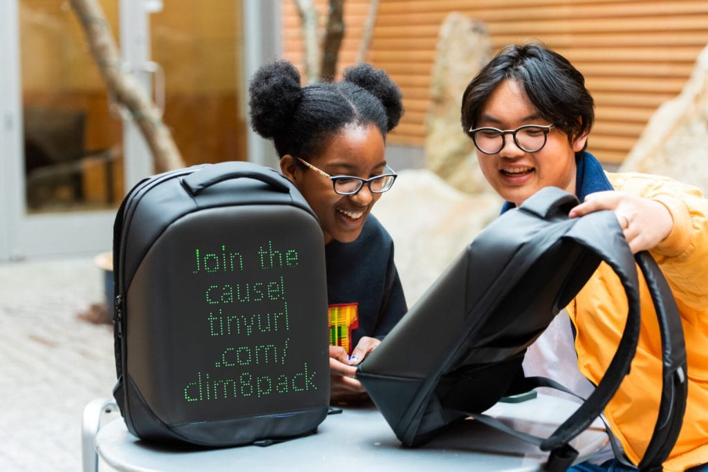 Two students sit in a table with a backpack in front of them that reads "Join the cause! tinyurl.com/clim8pack"
